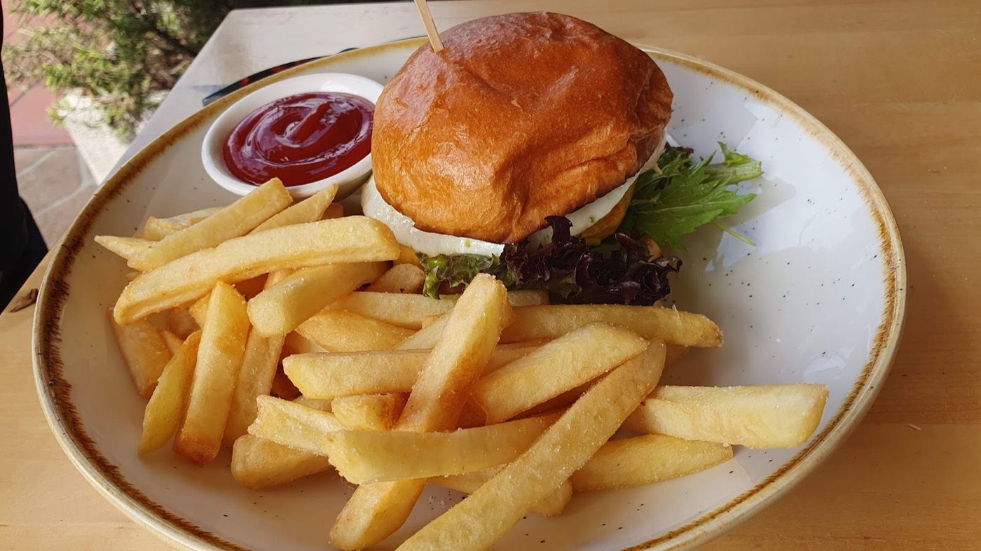 A burger in a glossy bun with a pot of tomato sauce and a pile of chips on a speckled plate