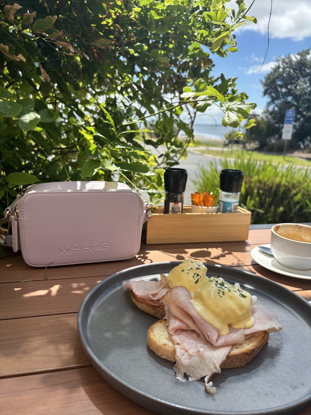 Eggs benedict with ham and hollandaise on sourdough, on a grey plate at an outdoor table with a coffee, greenery and the sea behind