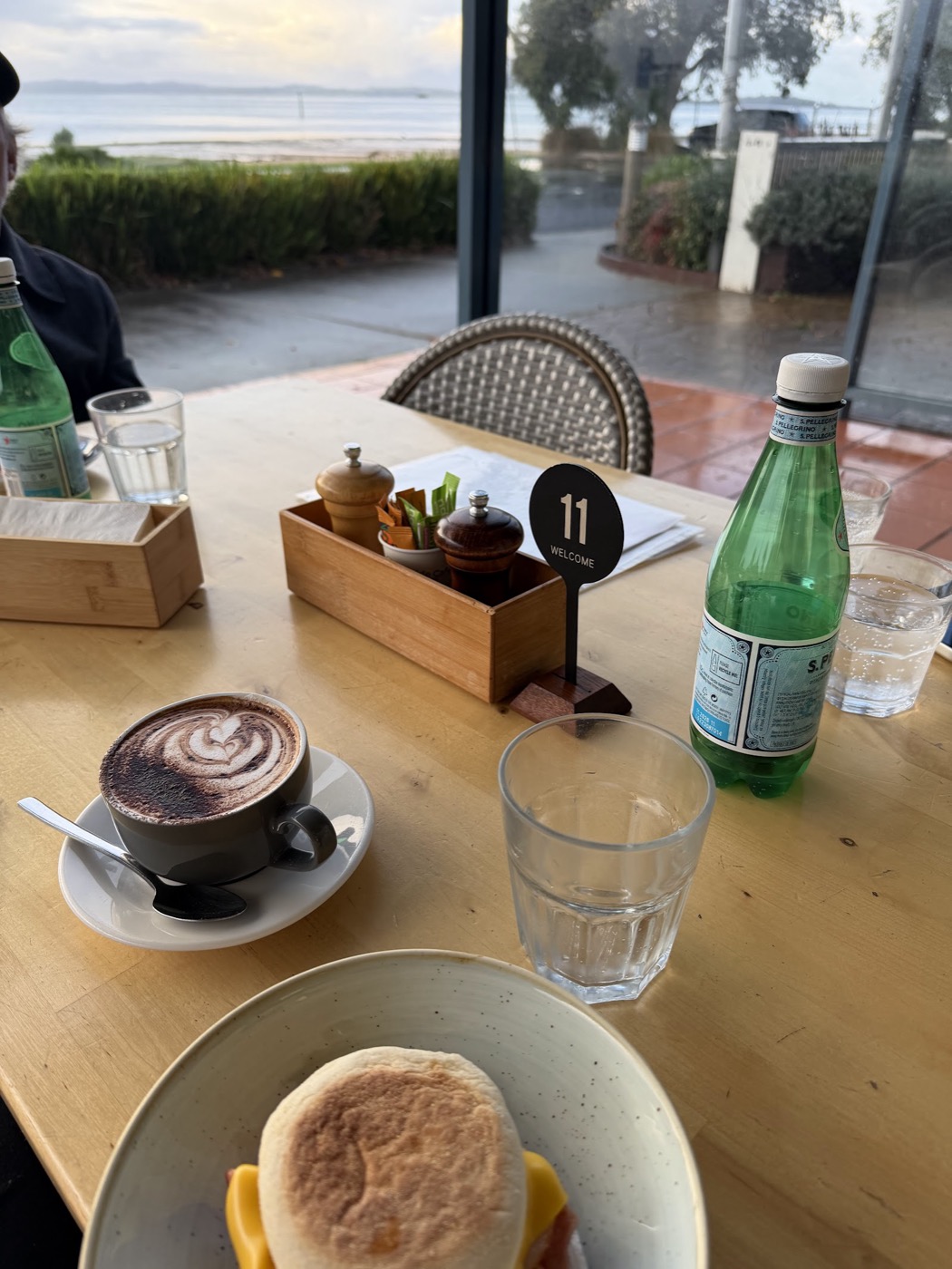 A timber table by the window with a coffee, a muffin on a plate, condiments and a green bottle of water